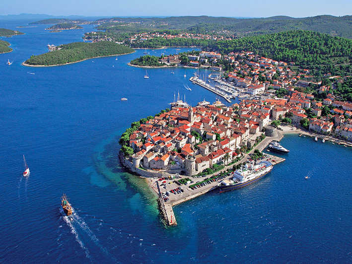 Aerial shot of a coastal town with deep blue water surrounding it and deep green forest in the distance